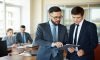 Confident businessman pointing at document while explaining his idea to his partner on background of their colleagues Paperwork